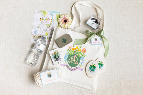 A wedding welcome bag by The Lux Box Co with a floral design contains a map, water bottle, pink flower, tin box, snacks, and cookies. Ideal for gifts for social events, the bag showcases initials and date for that personalized touch.