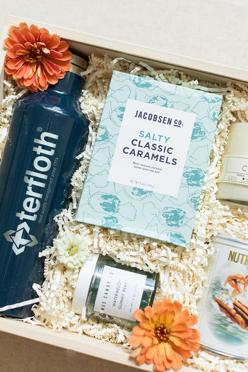 Gift box with a blue water bottle, salty classic caramels, candle, small jar, tin, and orange flowers on white paper shreds.