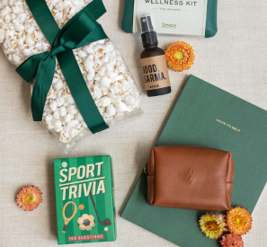 Flat lay of a corporate gift set by The Lux Box Co, featuring popcorn with a green ribbon, wellness kit, room spray, sport trivia cards, a leather pouch, a green notebook, and small flowers on a beige background.