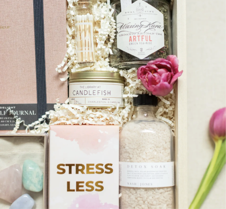 Gift box with a journal, candle, tea, bath salts, crystals, and a Stress Less card. Pink tulip nearby.