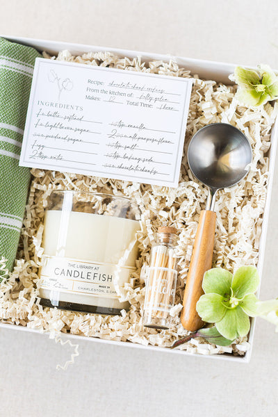 Discover our captivating gift box, perfect for corporate gifting or social events. It features a green kitchen towel, a recipe card, a candle, a small bottle, and a metal scoop with a wooden handle nestled among delicate green flowers on shredded paper packaging.