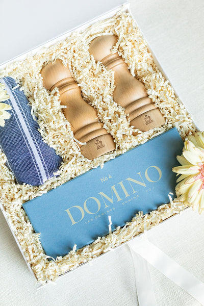 Gift box with two wooden pepper mills, a blue and white striped cloth, and a blue Domino booklet, surrounded by decorative shredded paper and flowers.