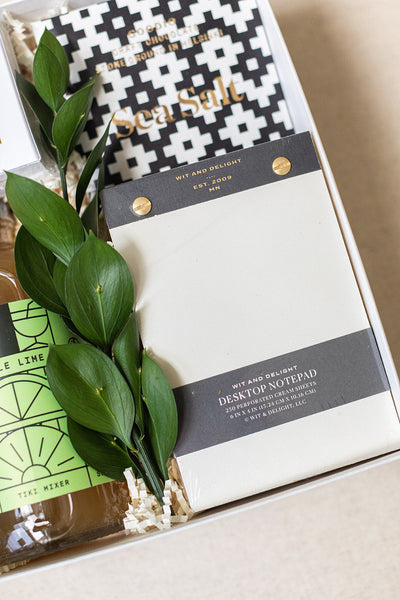 Stylish gift box with a desktop notepad, chic patterned item, Sea Salt chocolate bar, green foliage, and a Lime Tiki Mixer bottle nestled in straw.
