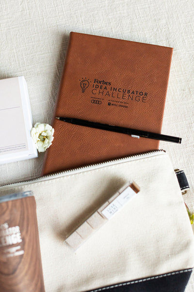 A notebook labeled "Forbes Idea Incubator Challenge," a pen, a small white flower, and a pouch are arranged on a light surface—an ideal ensemble for corporate gifting.