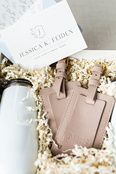 Gift box containing two beige leather luggage tags, a white insulated tumbler, and a card with the name Jessica K. Feiden among shredded paper filler.