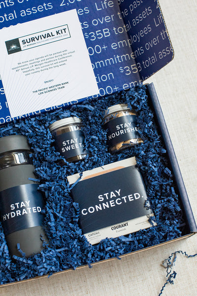 Open gift box labeled Survival Kit contains a bottle, two jars, and a box. Items are labeled Stay Hydrated, Stay Sweet, Stay Nourished, and Stay Connected, surrounded by blue packaging.