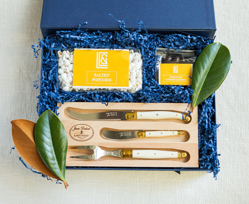 A gift set in an open navy blue box with blue shredded paper filler, featuring a wooden tray holding three white-handled metal knives and a matching fork. Inside the box are a bag of salted popcorn, a small packet of chocolate-covered appetizers, and branded yellow labels.