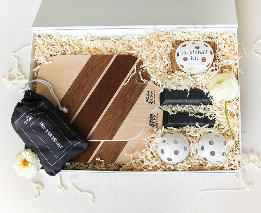 A wooden gift box filled with shredded paper packaging, containing a striped wooden paddle, a black drawstring pouch labeled 'Pickleball Kit', a round 'Pickleball Kit' tin, two white pickleball balls, and two black paddle grips. Decorative white flowers are placed around the box on a light beige surface.