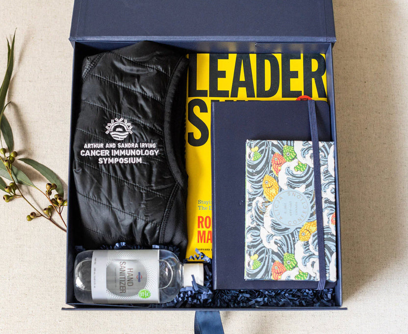 An open navy gift box arranged with a black quilted jacket printed with 'Arthur and Sandra Living Cancer Immunology Symposium,' a yellow book with bold black lettering, a patterned notebook, and a small bottle of water, all on a beige surface.