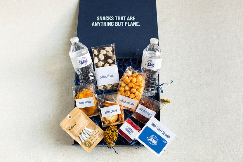 A curated snack gift box set on a light beige fabric background, featuring AMM branded water bottles, packaged chocolate bar, popcorn, chips, dried fruit, and a blue gift card, arranged in blue packing paper with a navy branded box behind.