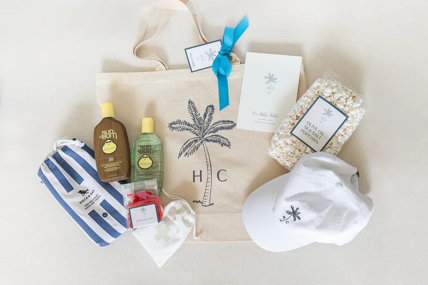 A curated gift set arranged on a light beige surface, featuring a beige canvas tote bag with a black palm tree and 'HC' monogram, a blue-and-white striped pouch, two small bottles, a white cap with a black logo, a bag of popcorn, and a white card tied with a blue ribbon.