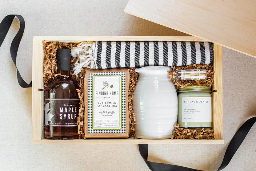 A wooden gift box filled with shredded paper and a black-and-white striped towel, arranged with a bottle of maple syrup, a box of buttermilk pancake mix, a white ceramic pitcher, and a candle.