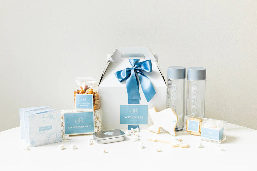 A curated gift set arranged on a white surface against a light neutral background. The set includes a white gift box with a blue ribbon, two clear bottles with gray caps, several small boxes and packets, a small tin, and a white animal-shaped item, all coordinated in pale blue, white, and gray tones.