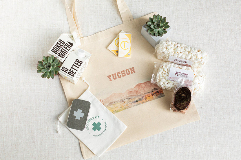 A beige canvas recovery tote bag laid flat on a light fabric surface, surrounded by gift items including boxed water, a small pouch with a green medical-style cross, bags of popcorn, a chocolate-covered stick, and two small succulent plants. The bag has the word TUCSON printed in muted orange-brown across the front.