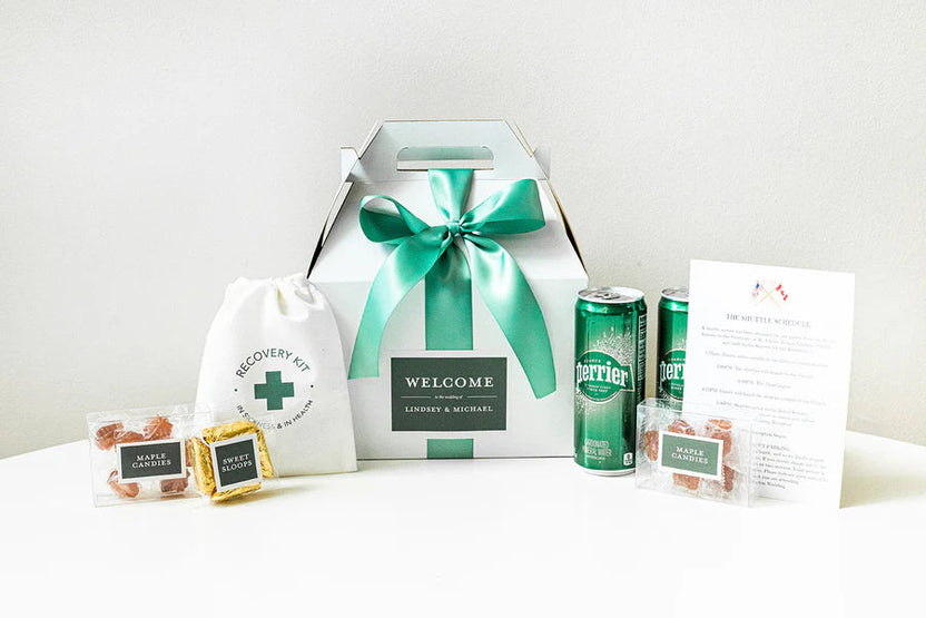 A curated gift set arranged on a white surface against a light neutral wall. The set includes a white gift box with a large teal ribbon and a front label reading 'WELCOME' and 'LINDSEY & MICHAEL,' a white drawstring pouch with a green cross and 'RECOVERY KIT' text, two green Perrier cans, two small packaged snacks, and a printed card.