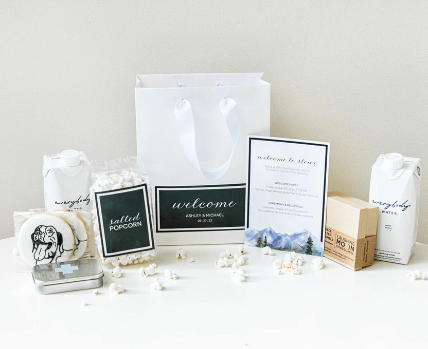 A wedding gift basket arrangement featuring a white paper gift bag, a custom 'welcome' sign, a mountain-themed invitation, a tin of cookies, bags of popcorn, a box of water, and a small card, styled on a white surface against a light beige background.
