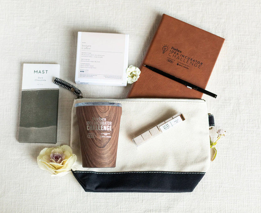 A styled flat-lay of a cream canvas zipper pouch with a navy bottom trim, containing a wooden-textured travel mug and a white pen. Surrounding the pouch are a brown leather notebook, a gray card, a white notepad, and small white flowers on a textured off-white fabric background.