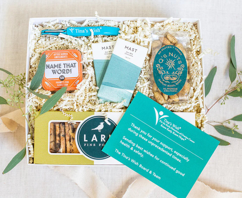 A curated gift box from Tina's Wish, filled with shredded paper and containing a blue bandana, a teal and orange 'Name That Word!' game card, two white MAST socks, a tin of Nutra Gold crackers, a box of Lara Pine Fingers, and a teal gift message card, styled with eucalyptus on a light fabric background.