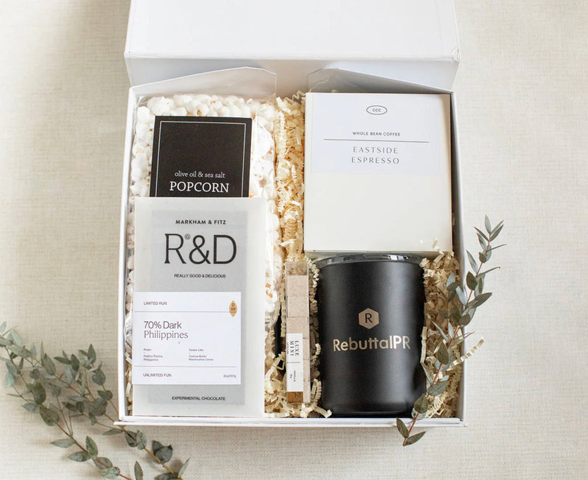 A curated gift box set containing a R&D coffee bag, a marble and stone popcorn package, a black Rebuffal PR candle, and a white espresso coffee bag, arranged in white crinkle paper inside a white box on a beige linen background with decorative greenery.