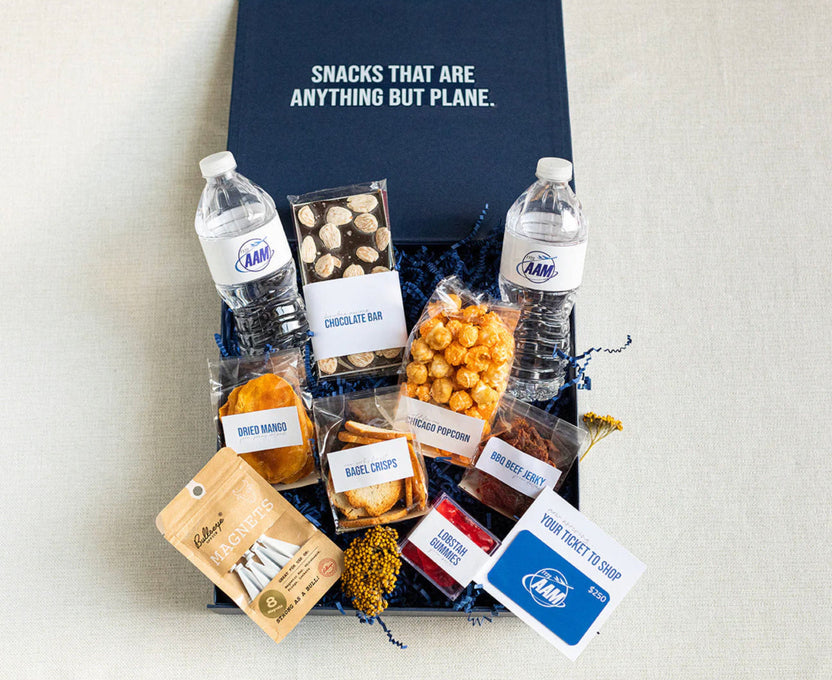 A curated snack gift box from Ram's Club, featuring an assortment of packaged snacks, two water bottles, and a blue gift box with the slogan 'SNACKS THAT ARE ANYTHING BUT PLANE.' on a light fabric background.