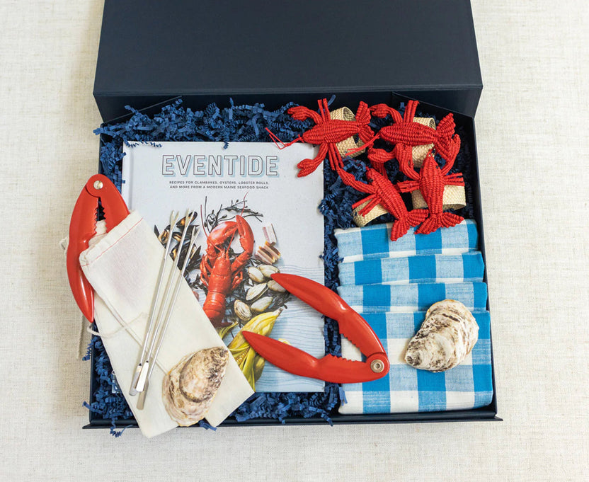 A curated seafood gift box set displayed in a black lid, filled with blue crinkle paper. The set includes a 'Eventide' magazine, a white napkin with red crab claws, a blue-and-white checkered napkin with a lobster shell, red rope crab decorations, and red crab claws.