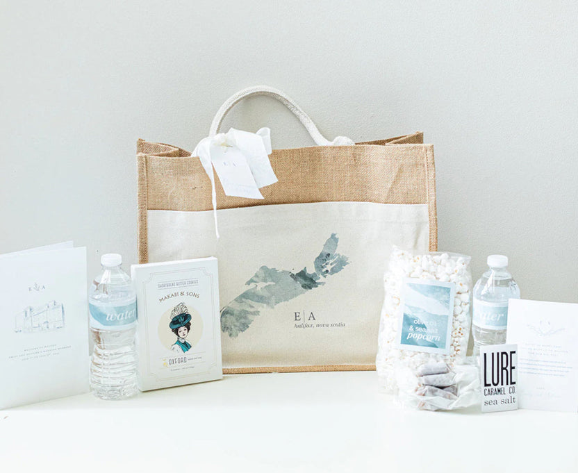 A gift set arranged on a white surface against a light gray wall, featuring a natural jute tote bag with a cream front panel printed with a gray map design and the initials 'E.A', along with water bottles, printed cards, and a bag of marshmallows.