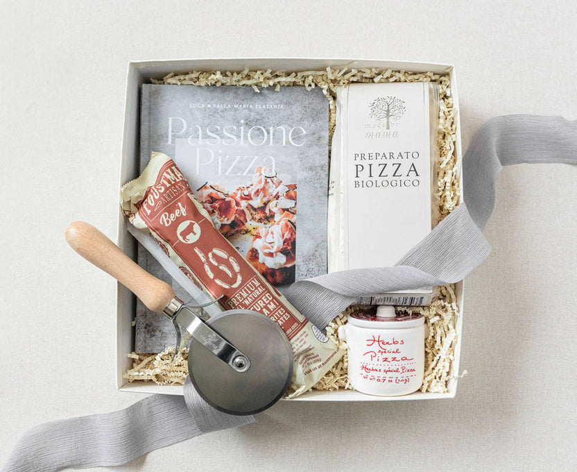 A pizza-themed gift box set containing a pizza cutter, a book titled 'Passione Pizza', a package of 'Bella+Ciao' pizza dough, a small jar of sauce, and a white box of 'Preparato Pizza Biologico' bread, arranged in a white box with shredded paper fill and a gray ribbon.