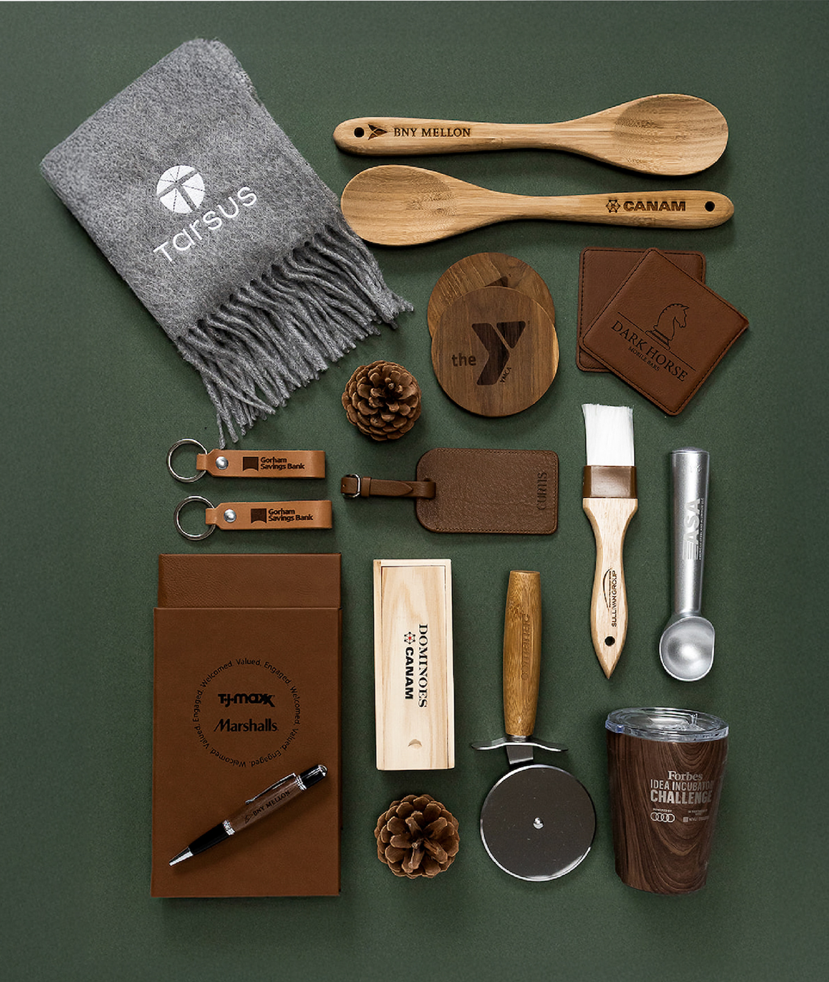 A curated flat lay of branded gift items on a dark green background, including a gray Totsus knit scarf, wooden spoons, engraved wooden coasters and tags, leather keychains, a brown notebook, a pen, a wooden box, a metal measuring cup, a small metal tin, pinecones, and a paintbrush.