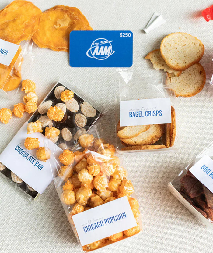 A gift basket assortment of snacks in clear bags with white labels, including Chicago Popcorn, Bagel Crisps, Chocolate Bar, and BBQ Bar, arranged on a light fabric surface with a blue AAM card above.