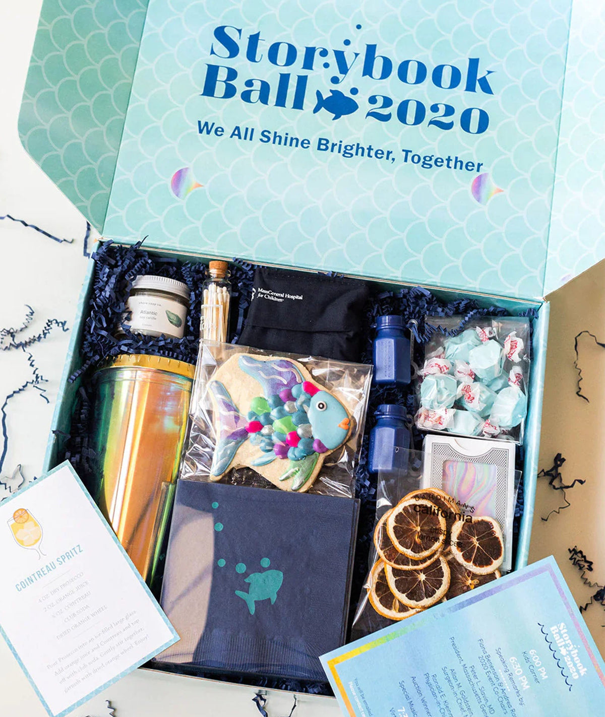 An open gift box labeled 'Storybook Ball 2020' filled with themed items, including a metallic tumbler, a fish-shaped cookie, wrapped candies, dried orange slices, a small bottle, and printed cards, photographed from above on a light surface.