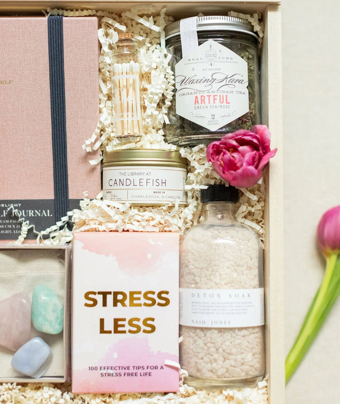 A curated gift box set filled with spa and self-care items, including a pink journal, a candle, bath salts, a match holder, crystals, and a pink flower, arranged in white shredded paper.