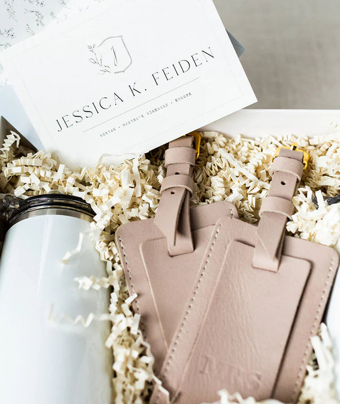 Two light blush pink leather luggage tags with gold-tone hardware nestled in white crinkle paper inside a gift box, with a white branded card reading Jessica K. Feiden visible at the top.