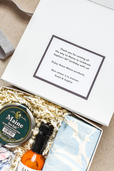 Discover a charming open gift box curated by The Lux Box Co, featuring Maine-themed delights: sea salt, hot sauce, a napkin, and a heartfelt note thanking guests for celebrating Sophie’s 50th birthday. Ideal gifts for social events that evoke fond memories.