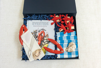 Gift box containing a cookbook titled Eventide, red lobster crackers, a checkered blue cloth, napkins, a shell, oyster forks, and a rope crab decoration in decorative shredded paper.