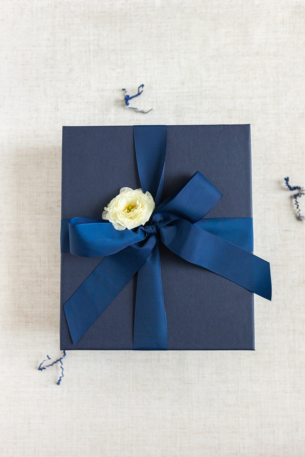 A dark blue LUX BOX co. gift box, adorned with a matching ribbon and white flower, rests elegantly against a light fabric background, ideal for holding the Duckbill - Teacher Appreciation Gift or gel tip pens.