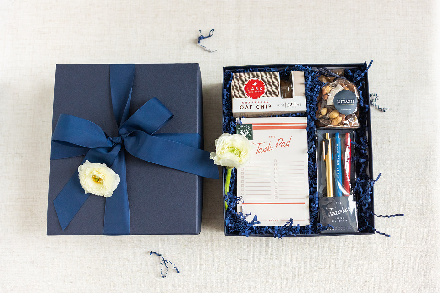 The LUX BOX co. Duckbill Teacher Appreciation Gift is beautifully wrapped with a navy ribbon and includes cranberry oat cookies, graham crackers, gel tip pens with a task pad, and a dark chocolate bar. Its elegantly adorned with white flowers and nestled in blue shredded paper.