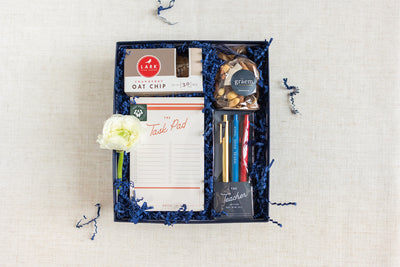 The LUX BOX co. Duckbill Teacher Appreciation Gift features a premium notebook, gel tip pens, dark chocolate bar, almonds, oat chip cookies, and a delicate white flower arranged on blue shredded paper.