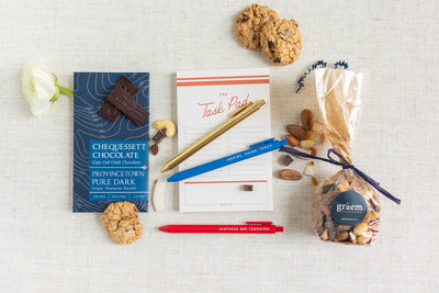 Give a thoughtful gift with the Duckbill Teacher Appreciation Gift from LUX BOX co., featuring gel tip pens, cookies, a dark chocolate bar, pencils, a notepad, and a bag of nuts elegantly arranged on a light background with a single white flower.