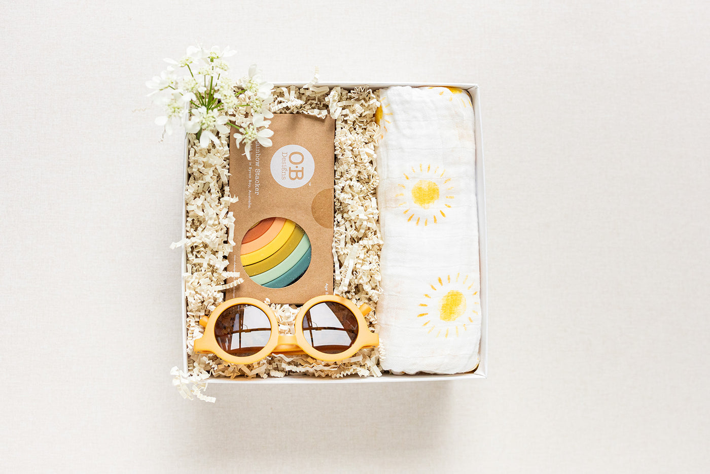The Duckbill New Baby Gift by LUX BOX co. includes a sun-patterned cotton swaddle, sunglasses, and a silicone rainbow stacking toy, adorned with decorative white flowers and nestled in shredded paper - an ideal baby gift.