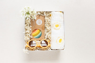 The Duckbill New Baby Gift by LUX BOX co. includes a sun-patterned cotton swaddle, sunglasses, and a silicone rainbow stacking toy, adorned with decorative white flowers and nestled in shredded paper - an ideal baby gift.
