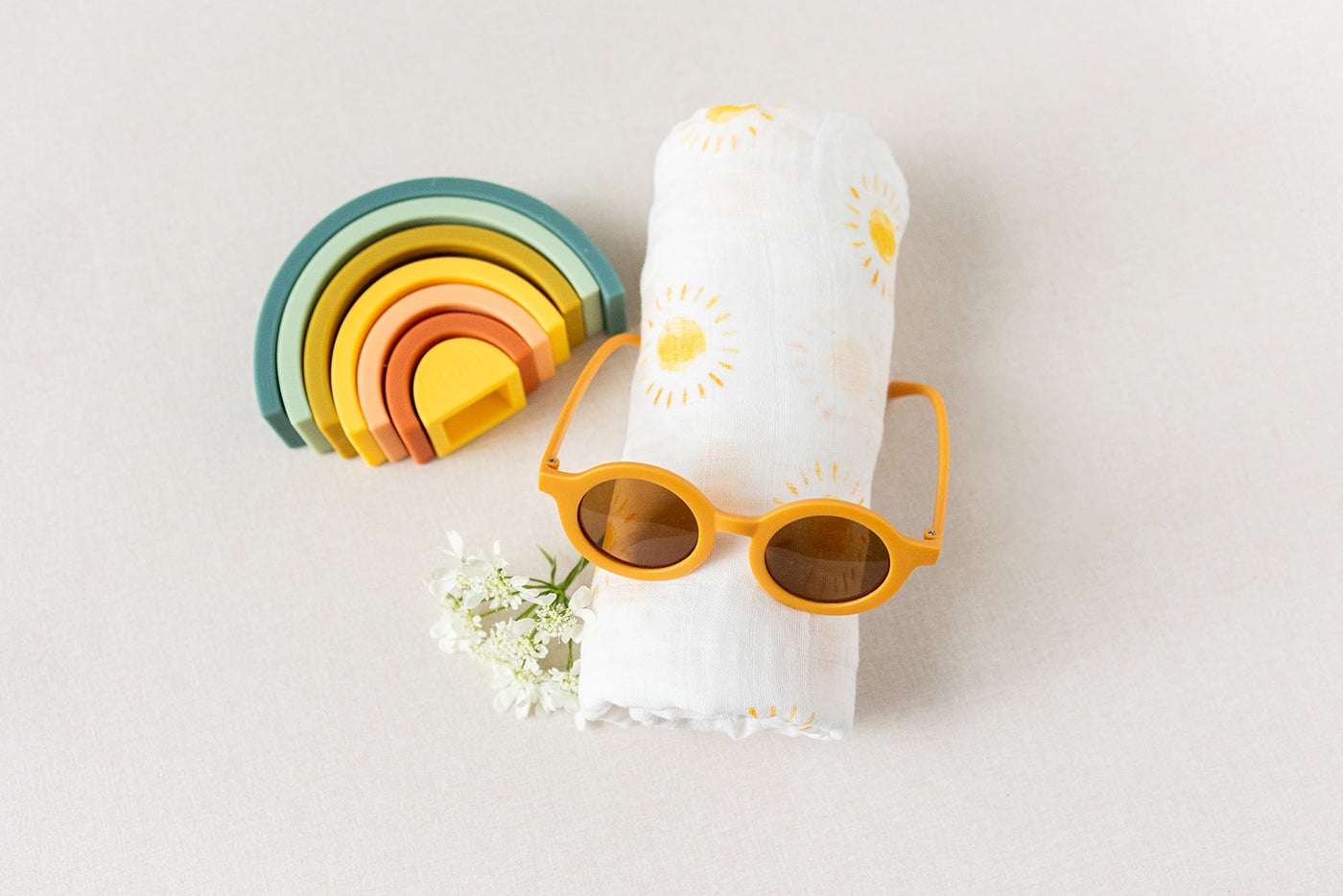 The Duckbill - New Baby Gift by LUX BOX co. includes a folded white cotton swaddle with sun patterns, yellow sunglasses, a multicolored silicone rainbow stacking toy, and delicate white flowers on a light surface for a charming presentation.