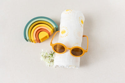 The Duckbill - New Baby Gift by LUX BOX co. includes a folded white cotton swaddle with sun patterns, yellow sunglasses, a multicolored silicone rainbow stacking toy, and delicate white flowers on a light surface for a charming presentation.