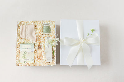 The Duckbill - Thinking of You by LUX BOX co. features a white gift box with a bow, containing cashmere socks, a soothing candle, luxurious hand lotion, and delicate dried flowers, all nestled in shredded paper.