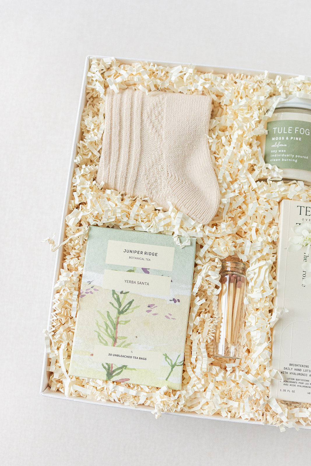 The Duckbill - Thinking of You gift box by LUX BOX co. includes cashmere socks, a scented candle, Yerba Santa tea, matches, hand lotion, and another delightful item all nestled in shredded paper.