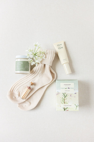 The Duckbill - Thinking of You by LUX BOX co. artfully arranges beige cashmere socks, a small jar of matches, Ten Over Ten hand cream, a Tule Fog candle, and Jasper Rose soap on a light surface with a small white flower—making it an elegant gift.