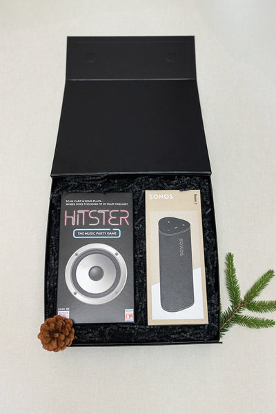 The LUX BOX co. Tune In & Play On features a Hitster music game and boxed Sonos portable speaker in a black gift box, accented with a pinecone and pine branch—ideal for energizing your next gathering.