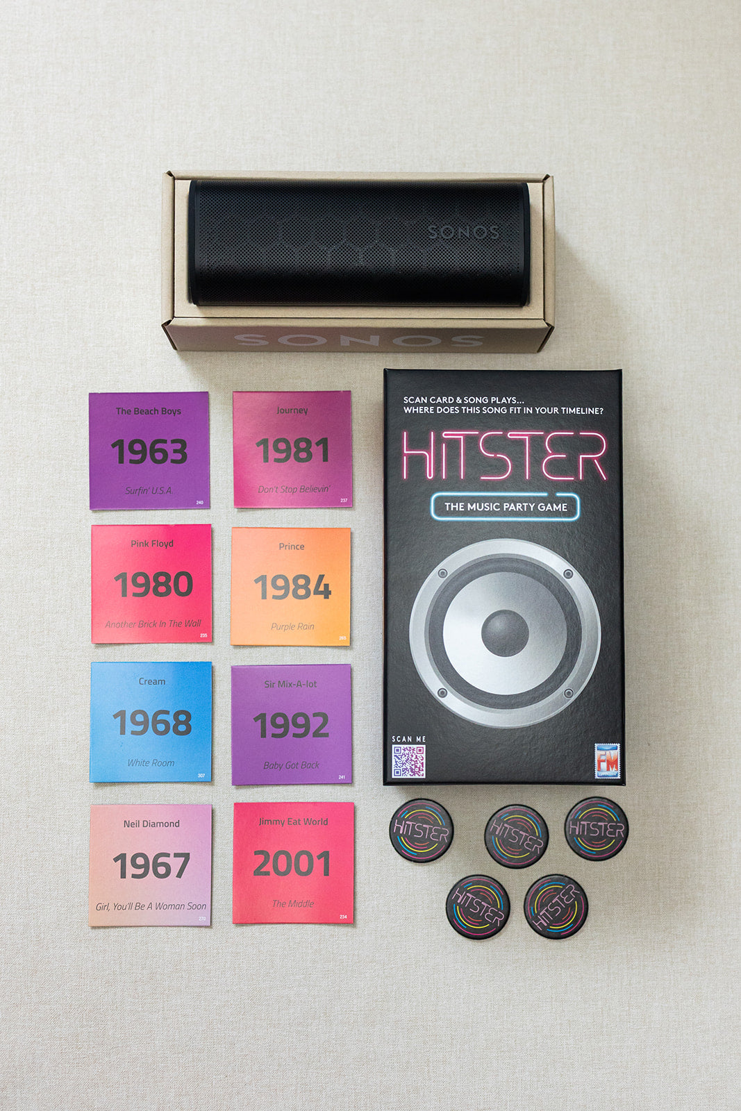 A LUX BOX co. Tune In & Play On set, featuring a music game box, four tokens, and eight vibrant genre cards with years and artists, is displayed on a light surface—ideal for your next song challenge.