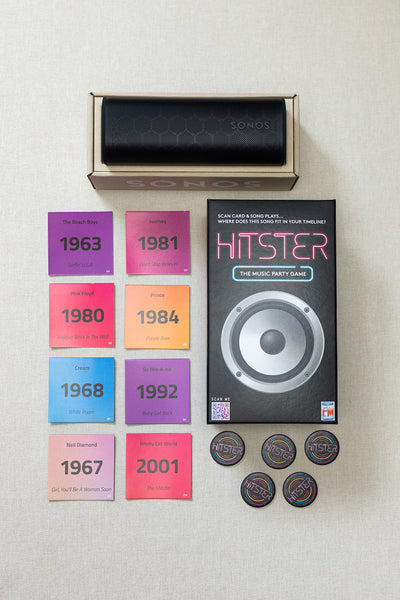 A LUX BOX co. Tune In & Play On set, featuring a music game box, four tokens, and eight vibrant genre cards with years and artists, is displayed on a light surface—ideal for your next song challenge.
