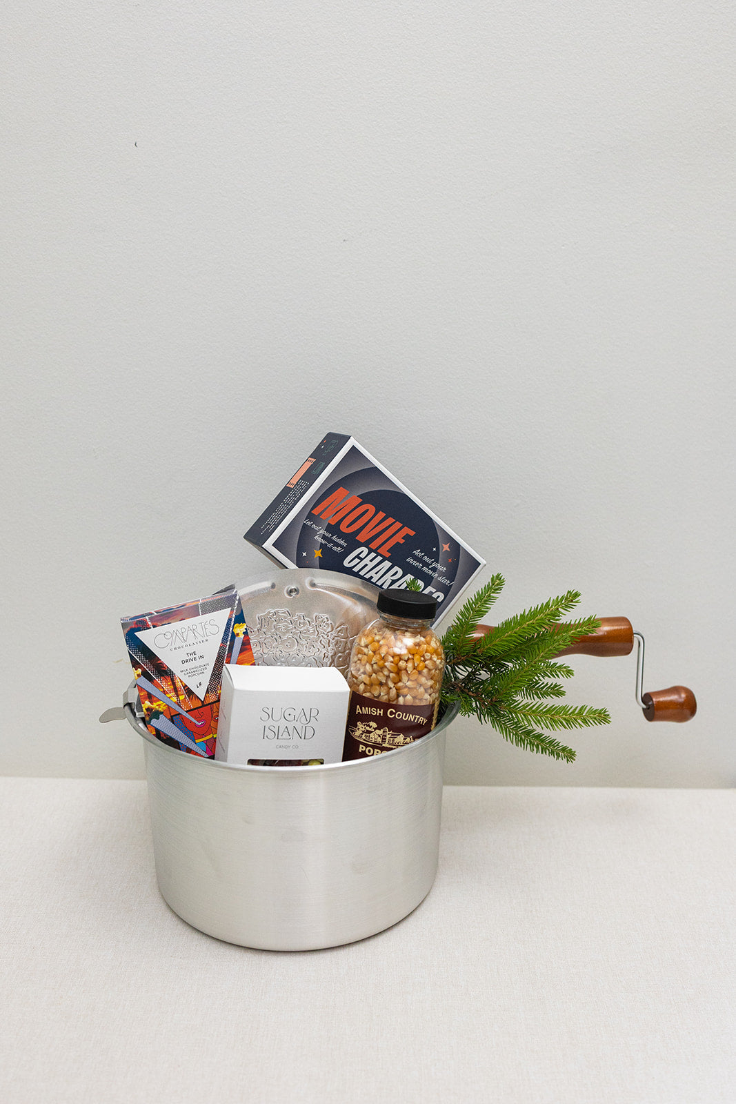 The Movie Night gift by LUX BOX co. features a silver pot with wooden handles filled with popcorn kernels, two chocolate bars, Movie Charades, and a pine branch—perfect for snack delights under the screen lights.
