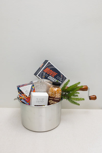 The Movie Night gift by LUX BOX co. features a silver pot with wooden handles filled with popcorn kernels, two chocolate bars, Movie Charades, and a pine branch—perfect for snack delights under the screen lights.